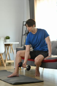 Millennial muscular male lifting dumbbell during morning workout at home. Fitness, weightlifting and bodybuilding concept.