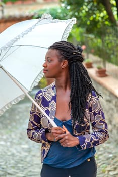 Portrait of an African woman with dreadlocks braid with umbrella parasol. Happy young woman feeling confident in her style. Fashionable woman standing in the street of old village against stone wall.