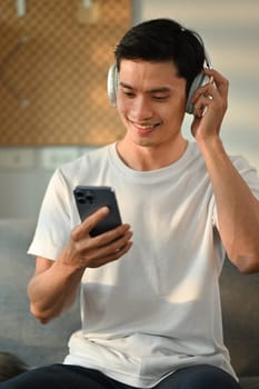 Joyful adult asian man enjoying listening to favorite music online on headphone and using smartphone in living room.