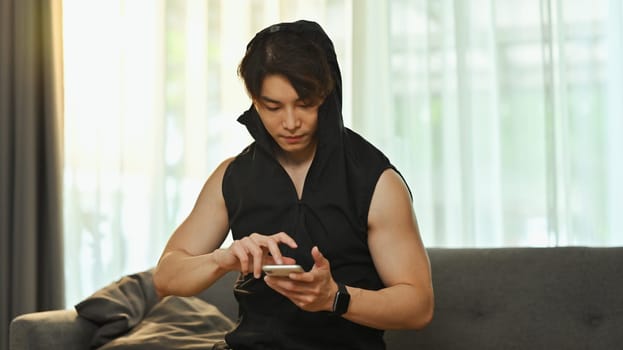 Young man in sportswear using smartphone, resting after working out at home. Fitness and healthy lifestyle concept.