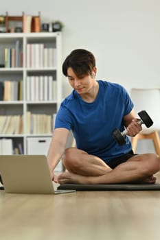 Muscular athletic man exercising with dumbbell and watching video online training on laptop during morning workout at home.