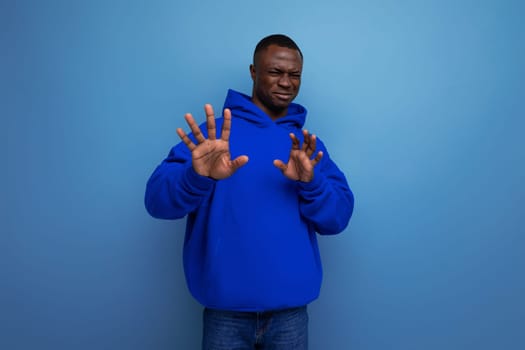 close-up of an emotional african young guy in a blue sweatshirt with a hood on a studio background with copy space.