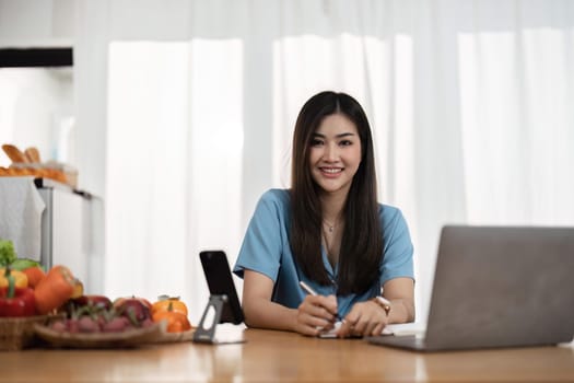 young woman is using laptop computer for remote work or studying online, she takes notes watching webinars or classes at kitchen table at home.