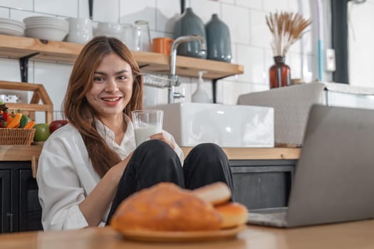 Beautiful young woman with laptop drinking milk in kitchen.