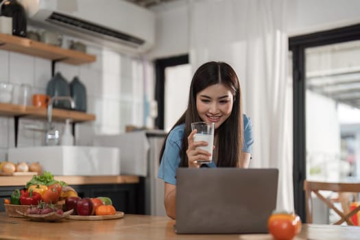 Beautiful young woman with laptop drinking milk in kitchen.