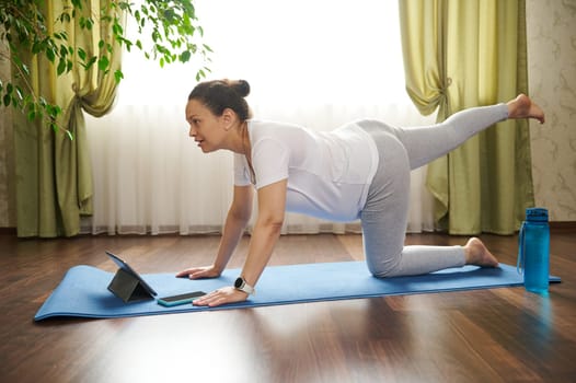 Young determined active pregnant woman watching online pregnancy yoga courses on digital tablet, doing prenatal sunbird, bird-dog pose, stretching her body on an exercise mat in cozy home interior