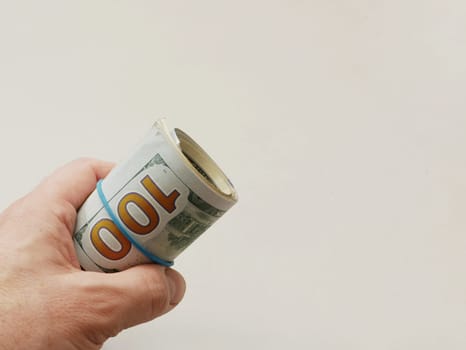 A man's hand holds a roll of 100 dollars bills with a blue rubber band on a white background.