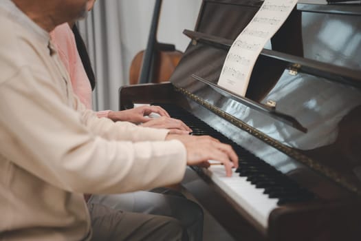 Family care. Happy daughter and elderly father playing music on the piano together in living room, Asian young woman teaching piano for senior man teaching at home, lifestyle life after retirement