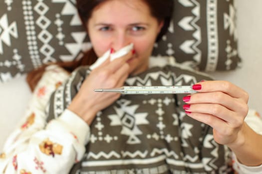 Ill woman laying in bed in her pyjamas, covered with blanket, holding napkin on her nose one hand, and mercury thermometer in other. Flu / grip sickness concept.