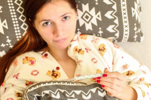 Top down view, blurred young woman laying in bed in her pyjama, holding thermometer (in focus). Grip / flu sickness concept.