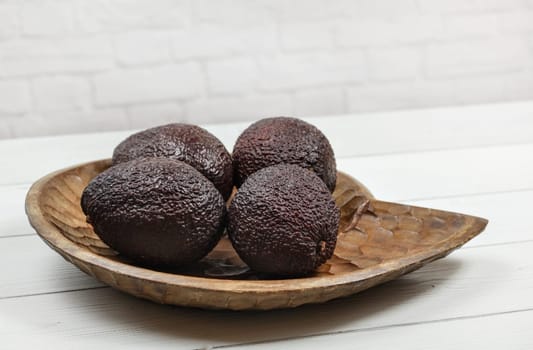 Four ripe dark brown avocados in wooden carved bowl on white boards desk.