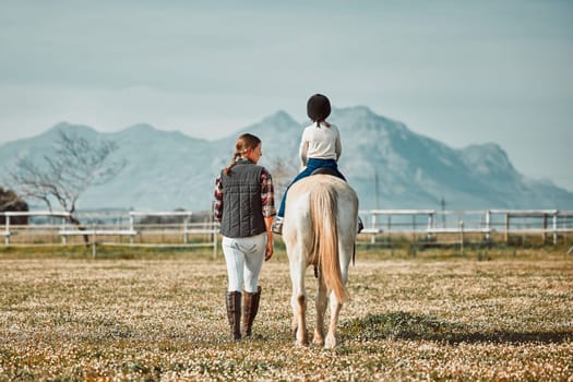 .Woman leading child on horse, ranch and mountain in background lady and animal walking on field from back. Countryside lifestyle, rural nature and farm animals, mom teaching kid to ride pony in USA