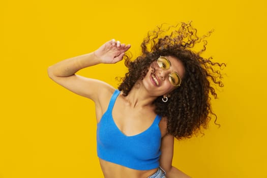 Woman with curly afro hair in a blue T-shirt on a yellow background dancing flying hair with sunglasses yellow, hand signs, look into the camera, smile with teeth and happiness, copy space. High quality photo