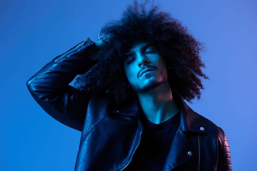 Portrait of fashion man with curly hair on blue background multinational, colored light, black leather jacket trend, modern concept. High quality photo