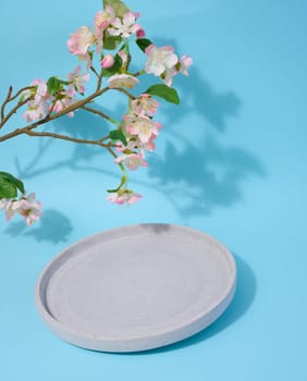 Round empty plate on a blue background and a cherry branch with flowers, a place to display cosmetics and products