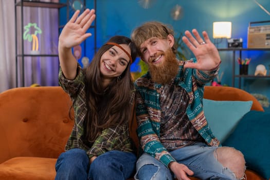 Happy young family marriage hippie couple man woman smiling friendly at camera and waving hands gesturing hello, hi, greeting or goodbye, welcoming at home. Husband and wife together on sofa in room