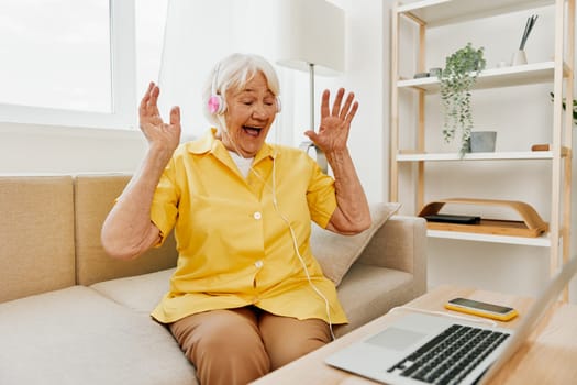 An elderly woman wearing headphones with a laptop communicating online by video call, sitting on the couch at home and working in a yellow shirt in front of a window, the lifestyle of a retired woman. High quality photo