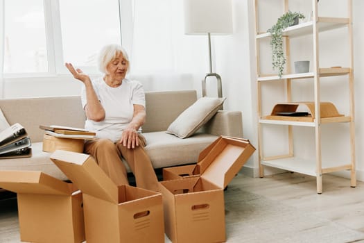 elderly woman sits on a sofa at home with boxes. collecting things with memories albums with photos and photo frames moving to a new place cleaning things and a happy smile. Lifestyle retirement. High quality photo