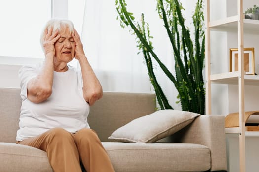 Elderly woman severe pain in the head sitting on the couch, health problems in old age, poor quality of life. Grandmother with gray hair holding her head, migraine and high blood pressure. High quality photo