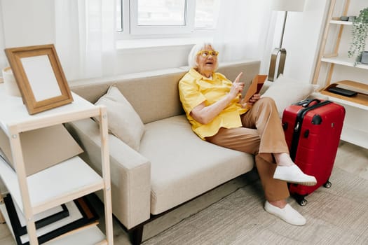 Happy senior woman with passport and travel ticket packed a red suitcase, vacation and health care. Smiling old woman joyfully sitting on the sofa before the trip raised her hands up in joy. High quality photo