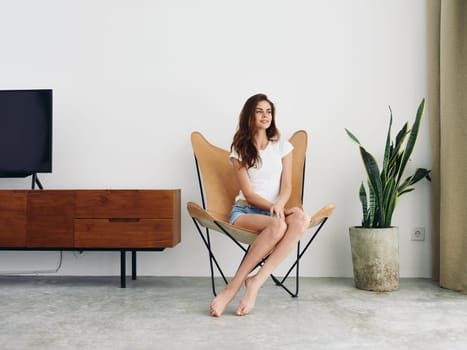 Woman sitting in leather armchair smile with teeth Lifestyle in white T-shirt and blue denim shorts, resting at home stylish modern interior with white walls, copy space. High quality photo