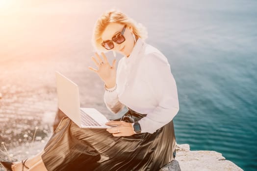 Digital nomad, Business woman working on laptop by the sea. Pretty lady typing on computer by the sea at sunset, makes a business transaction online from a distance. Freelance, remote work on vacation