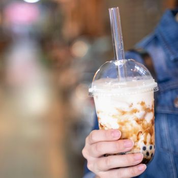 Young girl in denim jacket is drinking brown sugar flavored tapioca pearl bubble milk tea with glass straw in night market of Taiwan, close up, bokeh
