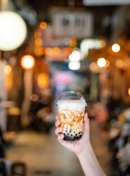 Young girl is holding and showing a cup of brown sugar flavored tapioca pearl bubble milk tea in night market of Taiwan background, close up, bokeh