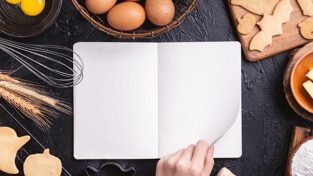Woman is reading cookbook recipe of making Halloween cookies with baking ingredients, design concept of cooking class, top view, flat lay, overhead.