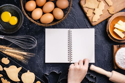 Woman is reading cookbook recipe of making Halloween cookies with baking ingredients, design concept of cooking class, top view, flat lay, overhead.