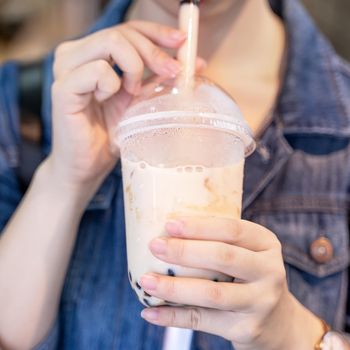 Young girl in denim jacket is drinking brown sugar flavored tapioca pearl bubble milk tea with glass straw in night market of Taiwan, close up, bokeh