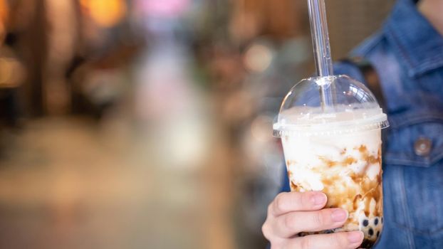 Young girl in denim jacket is drinking brown sugar flavored tapioca pearl bubble milk tea with glass straw in night market of Taiwan, close up, bokeh