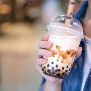 Young girl in denim jacket is drinking brown sugar flavored tapioca pearl bubble milk tea with glass straw in night market of Taiwan, close up, bokeh