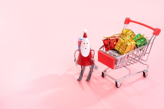 Christmas shopping concept, mini red shop cart trolley with Santa Claus toy and gift box isolated on pale pink background, blank copy space, close up