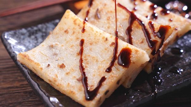 Pouring black soy sauce on ready-to-eat delicious turnip cake, Chinese traditional local dish radish cake in restaurant, close up, copy space.