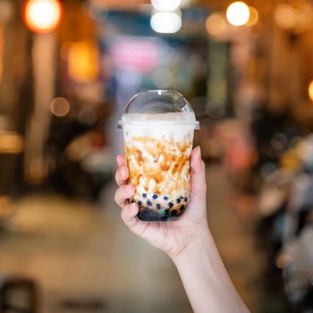 Young woman is holding, drinking brown sugar flavored tapioca pearl bubble milk tea with glass straw in night market of Taiwan, close up, bokeh