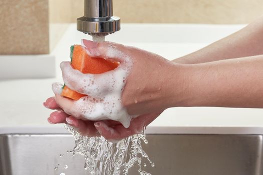 Women's hands in yellow protective glove with bottle of dishwashing liquid and sponge on kitchen. Cleaning concept.