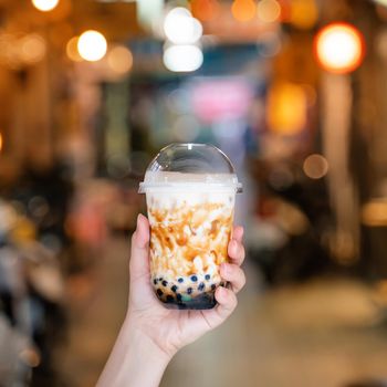 Young woman is holding, drinking brown sugar flavored tapioca pearl bubble milk tea with glass straw in night market of Taiwan, close up, bokeh