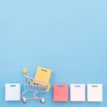 Abstract design element, annual sale, shopping season concept, mini yellow cart with colorful paper bag on pastel blue background, top view, flat lay