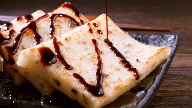 Pouring black soy sauce on ready-to-eat delicious turnip cake, Chinese traditional local dish radish cake in restaurant, close up, copy space.
