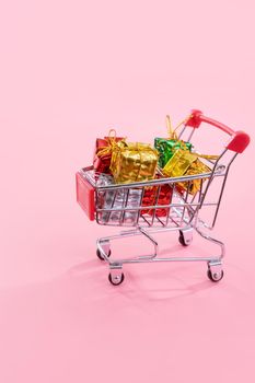 Annual sale, Christmas shopping season concept - mini red shop cart trolley full of gift box isolated on pale pink background, copy space, close up