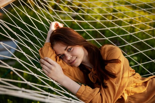 a happy woman is resting in a mesh hammock with her head resting on her hand, smiling happily at the camera with a smile, enjoying a warm day in the rays of the setting sun, lying in an orange dress. High quality photo