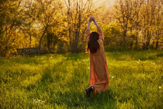 a slender woman with long hair stands in a field with her back to the camera, illuminated by the rays of the setting sun and joyfully raises her hands up. Horizontal photography on the theme of unity with nature. High quality photo