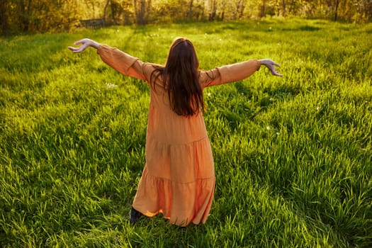 an attractive, slender, red-haired woman stands in a wide, green field during sunset in a long orange dress enjoying unity with nature and relaxation raising her arms to the sides while standing with her back to the camera. High quality photo