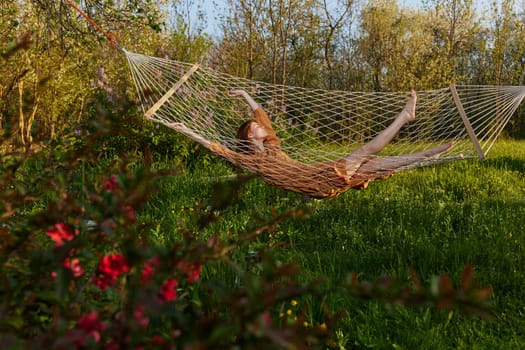 a happy woman in a long orange dress is relaxing in nature lying in a mesh hammock enjoying summer and vacation in the country surrounded by green foliage, happily lifting her legs. High quality photo