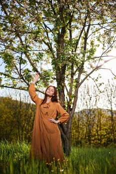 a sweet, attractive woman with long red hair is standing in the countryside near a flowering tree in a long orange dress and touching a branch with her hand looks at the camera. High quality photo