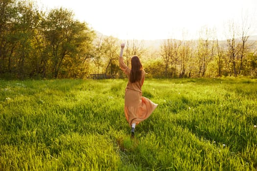 a relaxed slender woman enjoys the sunset standing in a green field with tall grass in an orange dress with her back to the camera, in warm summer weather. Horizontal photo. High quality photo