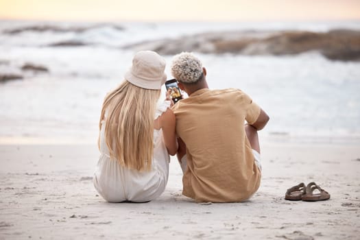 Rear view of a young couple taking a break and taking a selfie with a smartphone at the beach during sunset. Caucasian female and her mixed race boyfriend relaxing and enjoying the view at the beach.