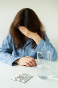 A young girl in pajamas sits at a table and holds her head against the background of a glass of water with pills. The concept of PMS, migraine, stress, depression