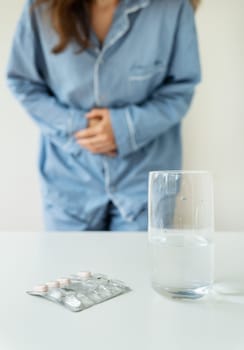 A young girl in pajamas stands and holds on to her stomach, there is a glass of water and pills on the table. The concept of PMS, indigestion, migraine, stress, depression